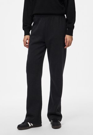 Personne portant un pantalon noir ample avec taille élastique, des baskets noires avec des rayures blanches et un haut noir à manches longues sur fond blanc.