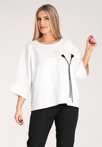 Biała, luźna bluzka z okrągłym dekoltem i krótkimi rękawami, z czarnymi akcentami kwiatowymi i sznurkiem z lewej strony. W połączeniu z czarnymi spodniami.