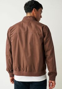 Chaqueta ligera marrón con textura suave, cinturilla y puños elásticos. Presenta un cuello clásico y un diseño minimalista.