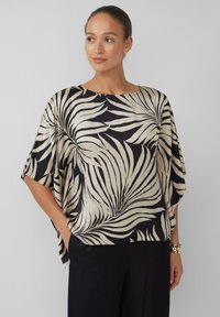 Vrouw in een losse zwart en beige top met abstracte bladpatronen en wijde mouwen, die met haar handen in haar zakken staat.