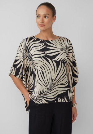 Femme portant un haut large noir et beige à motif de feuilles abstraites avec des manches larges et un pantalon noir, se tenant avec les mains dans les poches.