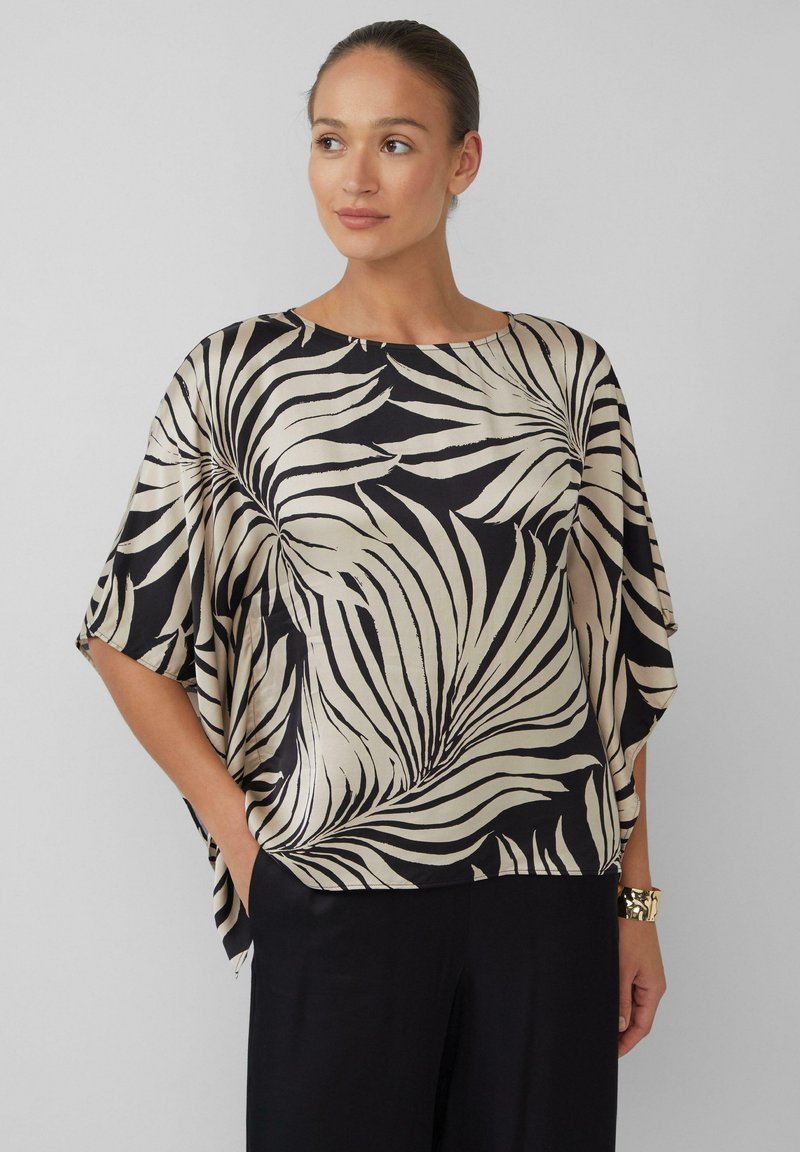 Vrouw in een losse zwart en beige top met abstracte bladpatronen en wijde mouwen, die met haar handen in haar zakken staat.