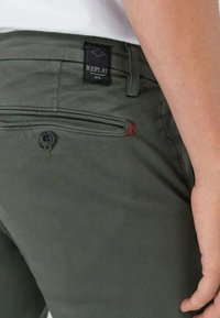 Pantalons vert olive avec une finition texturée, présentant une étiquette noire "REPLAY" et une petite couture rouge accent près de la poche arrière.