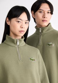 Sudaderas de color verde oliva con cremallera y cuellos altos. Presentan logotipos de cocodrilo bordados en el lado izquierdo del pecho. Tejido suave y texturizado.