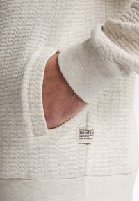 Teksturowany kremowy sweter z kieszenią i metką z napisem "Petrol Ind." Charakteryzuje się wzorem w zygzak oraz ściągaczami na mankietach.