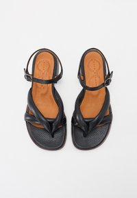 Sandalias de cuero negro con superficie texturizada, que cuentan con tiras anudadas y una plantilla de cuero marrón. Correa ajustable en el tobillo con hebilla.