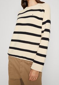 Maglione a righe beige e blu navy con collo tondo, texture a coste e un piccolo emblema sul petto. Maniche lunghe, vestibilità casual.