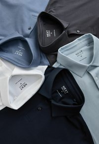 Seši salocīti vīriešu krekli ar apkaklīti melnā, baltā, gaiši zilā, tumši zilā un kaļķakmens pelēkā krāsā, izvietoti plakānā izkārtojumā.