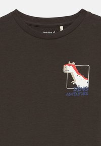 Dunkelbraunes Hemd mit einem kleinen Cartoon-Dinosaurier-Graphic und dem Text "Dino Abenteuer" im oberen Brustbereich.