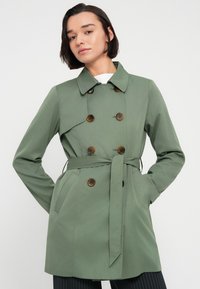 Πράσινο διπλόπατο παλτό από λείο ύφασμα, με καφέ κουμπιά, μέση με ζώνη και πλαϊνές τσέπες. Απλός γιακάς, μοντέρνος σχεδιασμός.