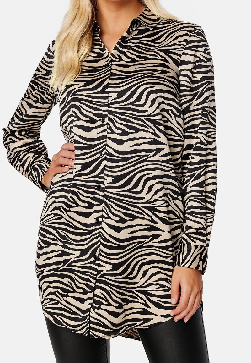 Femme portant une chemise à manches longues à imprimé zèbre noir et beige, boutonnée, avec un pantalon noir, debout avec une main sur la hanche.