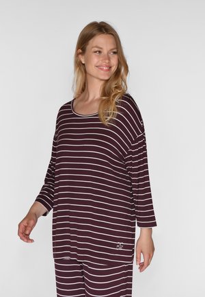 Lachende vrouw die een donkerpaarse en witte horizontaal gestreepte lange mouwen top en bijpassende broek draagt tegen een effen achtergrond.