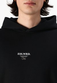 Svart hettegenser med snøring, med hvit tekst som sier "ESS.WRD." og ekstra merkevare under. Myk tekstur, enkelt design.