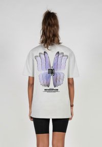 Gray short-sleeve t-shirt featuring a large purple butterfly graphic on the back, with black text below and a relaxed fit design.