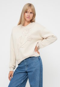 Crèmekleurige sweatshirt met bloemenborduursel in lichte draad. Voorzien van een ronde halslijn en geribbelde manchetten, gecombineerd met hoge blauwe jeans.