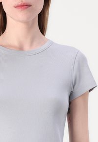 Šviesiai pilka, su gumbai siuvinėta trumpųjų rankovių marškinėliai su apvaliu kaklu, pagaminti iš glotnaus, tamprumo medžiagos. Pasižymi šiek tiek matoma tekstūra ir tvarkingais siūlais.