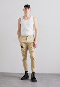 Pantaloni beige a sigaretta con dettagli testuali neri e chiusura con bottone. Indossati con una canotta bianca e stivali neri con lacci.