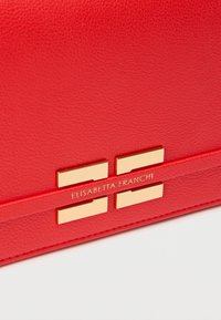 Κόκκινο δερμάτινο τσαντάκι χειρός με υφή, διακοσμημένο με πλακέτα λογότυπου σε χρυσό και μεταλλικά στοιχεία. Λεπτή σιλουέτα με σχεδιασμό ευθείας άκρης.