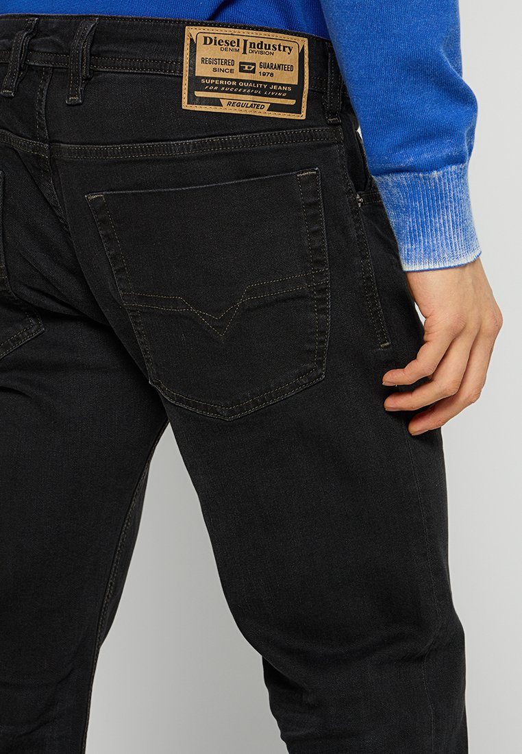 Jeans en denim noir avec une taille moyenne, présentant un design distinctif des poches arrière, un patch en cuir brun et des coutures jaunes contrastées.