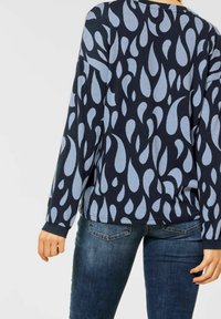 Femme portant un haut à manches longues bleu marine avec des motifs en forme de goutte d'eau bleu clair et un jean en denim bleu foncé, tournant le dos à l'appareil photo.