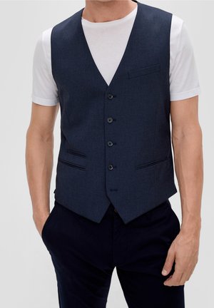 Man wearing a dark blue buttoned vest over a white short-sleeve t-shirt with dark pants, hands in pockets, against a plain background.