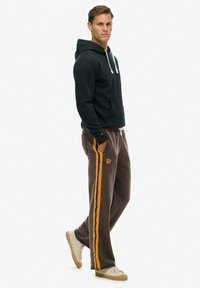 Sort hættejakke med snore, brune sweatpants med orange side-striber og logo, sammen med beige sneakers.