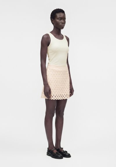 Γυναίκα που στέκεται, φοράει μπεζ αμάνικη μπλούζα, μπεζ φούστα από δαντέλα και μαύρα παπούτσια με αγκράφες, με απλό λευκό φόντο.