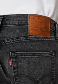 Svarte denimjeans med et brunt lærmerke som har gravert tekst, synlige sømmer, og en liten rød etikett på baklommen.