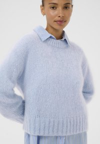 Pull tricot bleu clair avec une texture douce et duveteuse, col rond et ourlet côtelé, superposé à une chemise col rayée bleu clair et blanche.