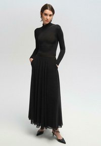 Top negro de manga larga con un corte ajustado, combinado con una falda maxi negra fluida que cuenta con bolsillos, finalizando con un dobladillo semitransparente. Zapatos de tacón negros.