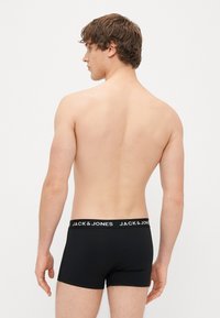 Črne bokserice z bela elastiko z blagovno znamko, izdelane iz gladkega materiala. Model je prikazan s hrbtne strani, kar razkriva prilegajoč dizajn.