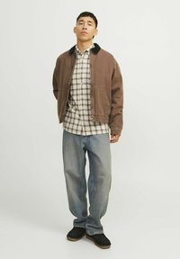 Chaqueta bomber marrón con un cuello negro, camisa de cuadros debajo, jeans holgados de lavados claros y calzado negro. Conjunto simple y casual.