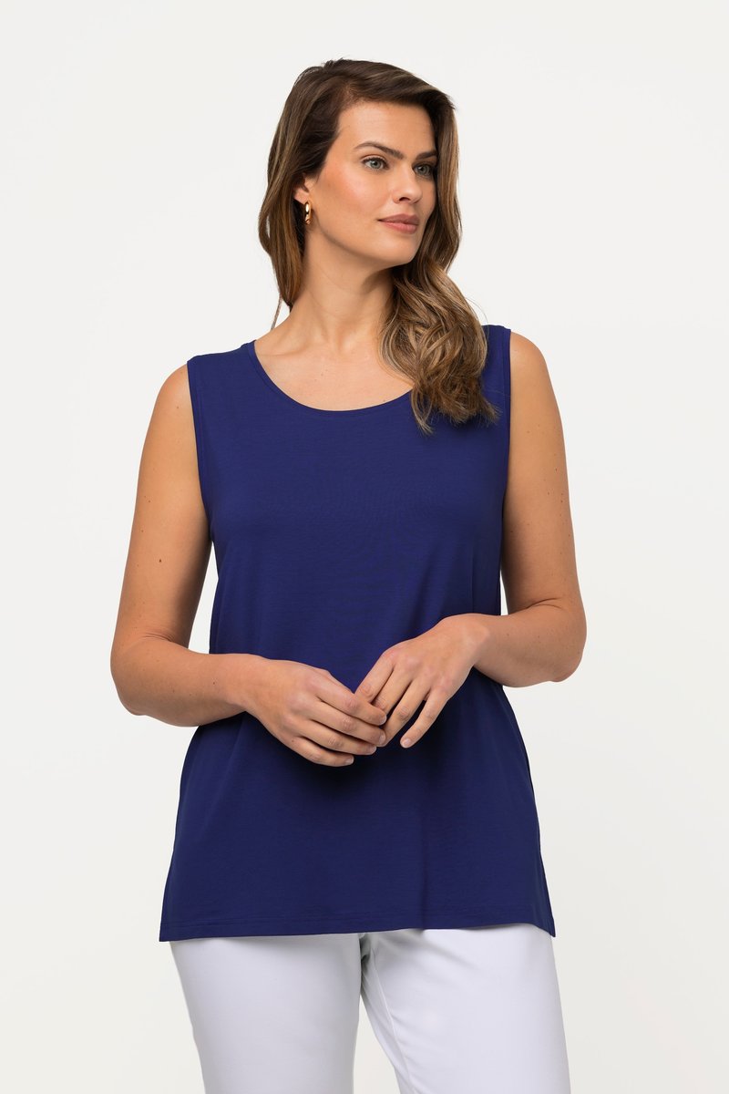 Donna con capelli castani mossi, che indossa orecchini a cerchio dorati, una canotta blu navy e pantaloni bianchi, in piedi con le mani sciolte incrociate.