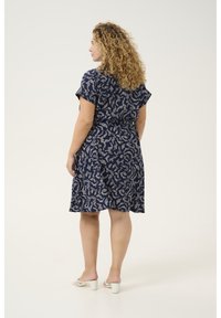 Navyblaues Kleid mit weißem abstraktem Muster, kurzen Ärmeln und einer taillierten Taille, die in einen knielangen Rock übergeht. Schlichte weiße Absatzsandalen.