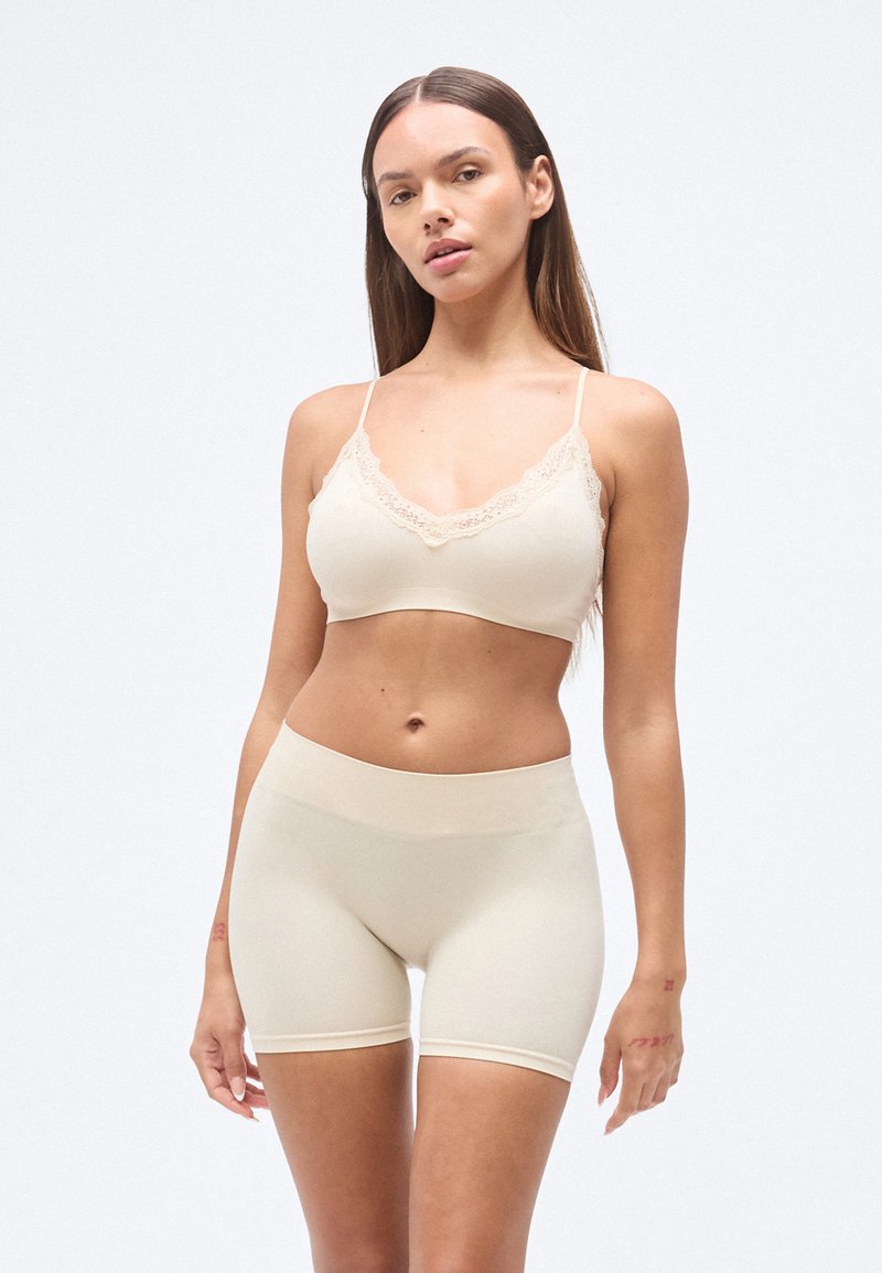 Brassière beige sans coutures avec bordure en dentelle et bretelles réglables, associée à un shorty beige taille haute, texture lisse, design ajusté.