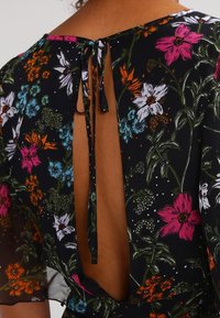 Musta kukkakuvioinen mekko, jossa on syvä selkädekoltee ja solmittava suljenta, koristeltu värikkäillä kukilla ja vihreillä lehdillä teksturoidulla kankaalla.