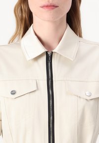 Beige jacka med spetsig krage, framdragkedja och två bröstfickor. Har kontrasterande svarta dragkedjor och glansiga knappdetaljer.