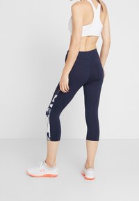 Leggings capri de color marino con bolsillo lateral, que presentan un patrón geométrico en azul y blanco. Fabricados con un material elástico, combinados con zapatillas deportivas blancas.