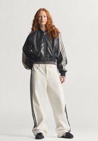 Kratka crna bomber jakna s teksturom krokodilske kože, bijele hlače visokog struka s crnim prugama sa strane, uparen s crnim cipelama.