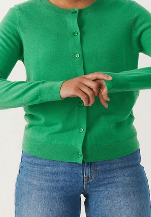 Person iført en lys grøn knappet cardigan og blå jeans, der justerer ærmet på venstre arm.