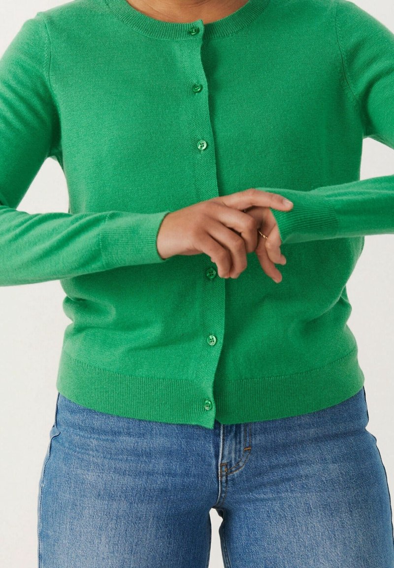 Personne portant un cardigan vert vif boutonné et un jean bleu ajustant la manche de son bras gauche.