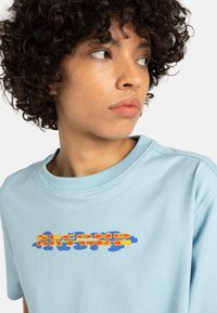 T-shirt en coton bleu clair avec un imprimé graphique qui indique "SECOND NATURE" en rouge et jaune vif, sur un fond bleu nuage.