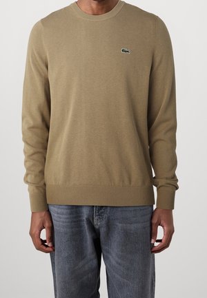 Mand iført beige crewneck-sweater med lille grønt krokodillelogo på brystet og blå jeans, stående mod en ensfarvet baggrund.