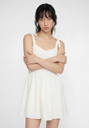 Jeune femme aux cheveux noirs portant une robe blanche sans manches avec des bretelles nouées aux épaules, bras croisés, sur un fond uni.