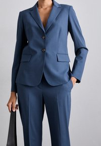 Blazer blu scuro su misura con due bottoni, rever a punta e tasche frontali, abbinato a pantaloni coordinati, tessuto liscio e design aderente.