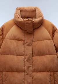Brązowa kurtka puchowa z wysokim kołnierzem z korduroy, z pikowanymi sekcjami i gładką powierzchnią. Tekstura jest zarówno miękka, jak i ciepła.
