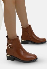 Botas de tobillo de imitación de cuero marrón con un bajo tacón cuadrado, cremallera lateral y acentos de hardware en tono dorado. Acabado suave con puntera redondeada.