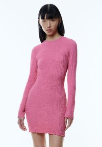 Kurzes rosa Kleid aus strukturiertem Stoff, mit langen Ärmeln und einem wellenförmigen Saum, eng anliegendem Schnitt und rundem Ausschnitt.