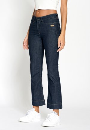 Vrouw draagt donkerblauwe rechtvallende jeans, wit cropped top en witte sneakers, staand voor een effen achtergrond.