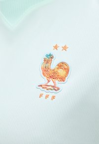 Zelta gailis emblēma ar divām zvaigznēm augšā un burtiem FFF apakšā uz gaismas zila teksturēta auduma.